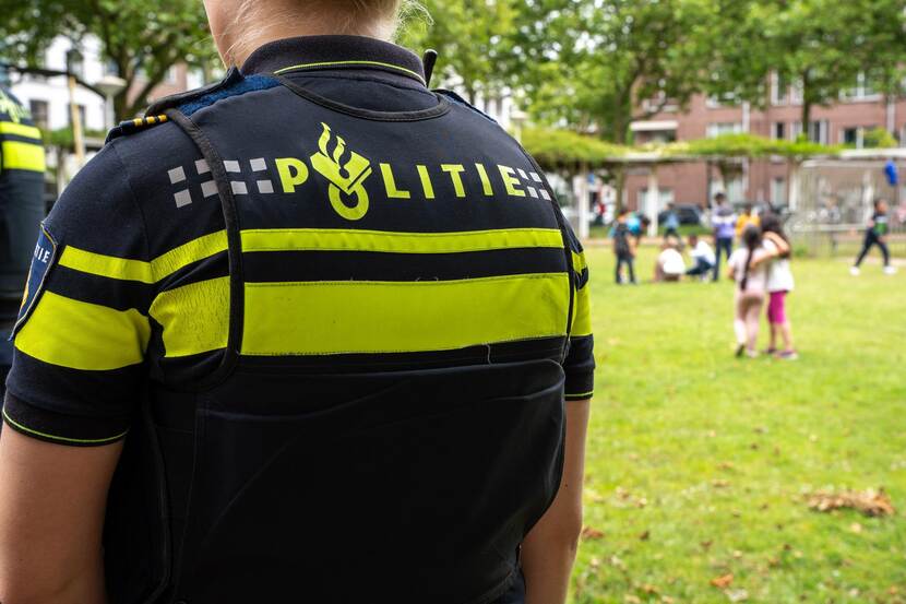 Agente in uniform in de wijk; gezien op de rug. De omgeving is een grasveld met spelende kinderen en op de achtergrond nieuwbouw woningen en bomen.