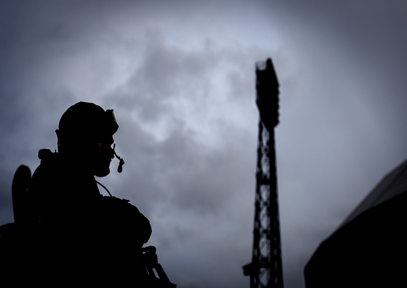 Silhouet van een militair of politieagent met helm, kogelwerend pak en een gedeelte van een machinegeweer bij een locatie waar ook het silhouet van een lichtmast te zien is. Dit alles tegen een dreigende bewolkte lucht.